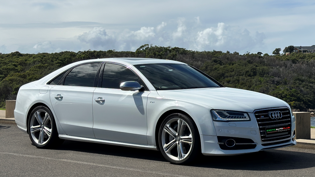 A white Audi S8 luxury sedan is parked on a paved area with scenic greenery and hills in the background. The car features sleek lines, tinted windows, and large alloy wheels, showcasing its sophisticated design and high-performance capabilities.