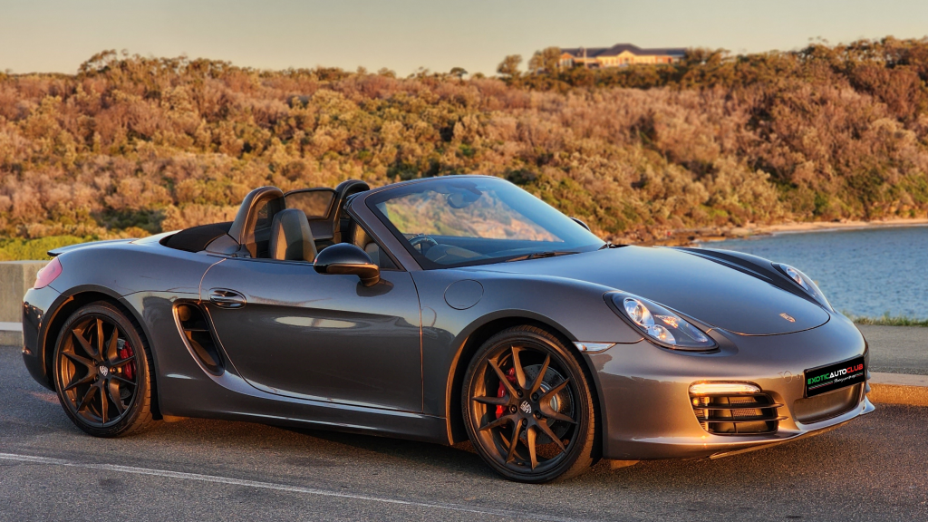 A sleek, gray Porsche Boxster convertible is parked on a road beside a serene body of water. The car's top is down, showcasing its luxurious interior. Surrounding the scene are trees and a view of a distant landscape under a clear sky at sunset.