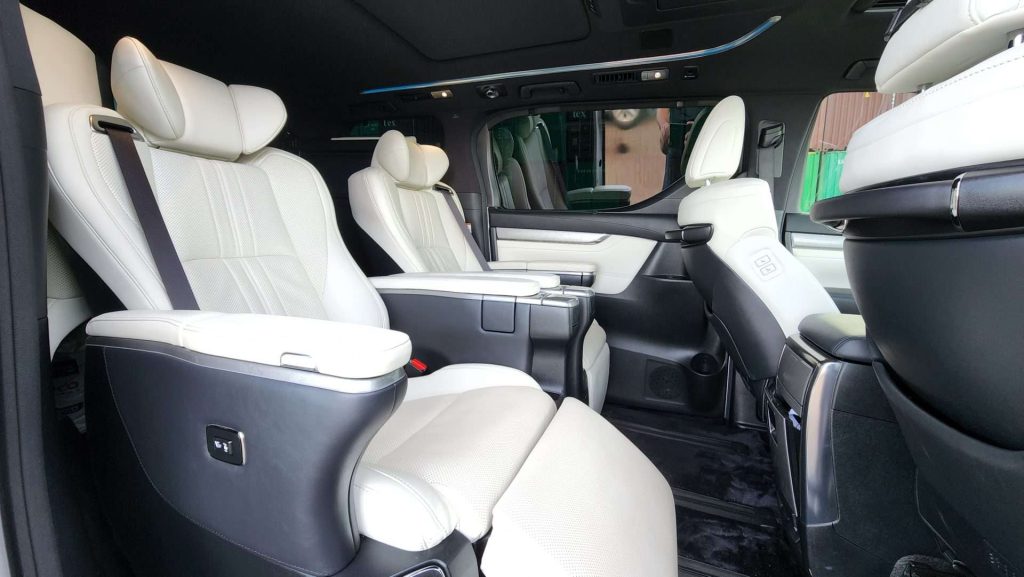 The image shows the interior of a luxury Alphard vehicle with spacious seating. The seats are upholstered in white leather with black accents. Captain's chairs in both the front and second row feature adjustable armrests, seat controls, and ample legroom, creating an executive lounge experience.