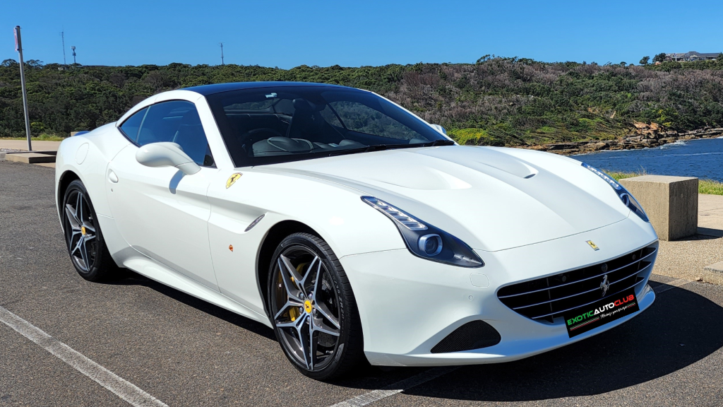 A white Ferrari California T parked on a paved area beside a scenic coastline with greenery and water visible in the background. The sleek curves and distinctive Ferrari logos of the sports car are highlighted as it sits near a beautiful coastal view under a clear blue sky.