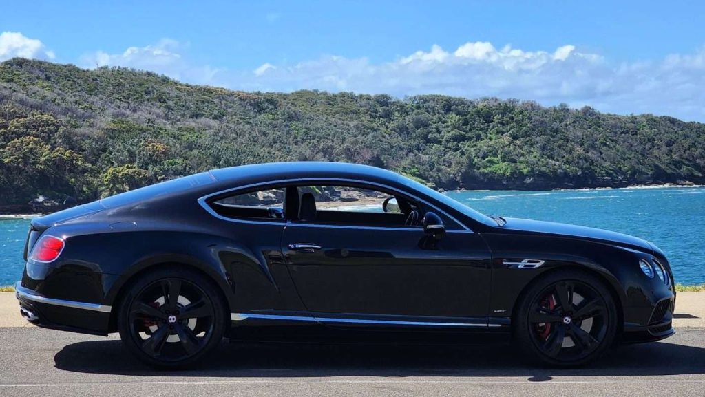 A sleek black Bentley Continental GT is parked near a scenic coastline. The ocean and green hillside provide a picturesque background under a partly cloudy blue sky. The car's polished exterior and sporty design, featuring its powerful V8s engine, are prominently showcased in the side profile image.