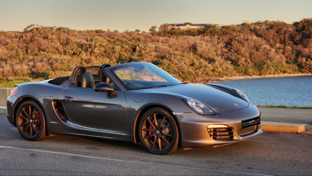 A sleek, dark gray Porsche Boxster convertible sports car is parked on the side of a road near a body of water. The top is down, and the car is positioned to show its side profile. In the background, there are trees and shrubs under a clear, sunlit sky.