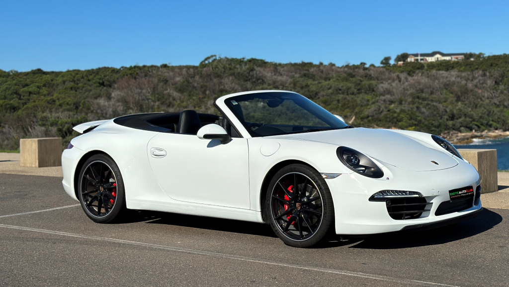 A white Porsche Carrera 911 convertible sports car is parked on a paved surface with a scenic background of greenery and a clear blue sky. The car's top is down, and it has sleek black wheels.