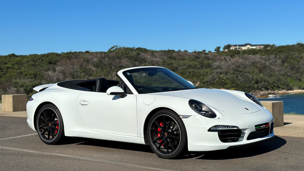 A white Porsche 911 Carrera S cabrio is parked in an outdoor setting with a clear blue sky. The car boasts a sleek, aerodynamic design with black wheels and red brake calipers. The background includes lush greenery and a glimpse of water.