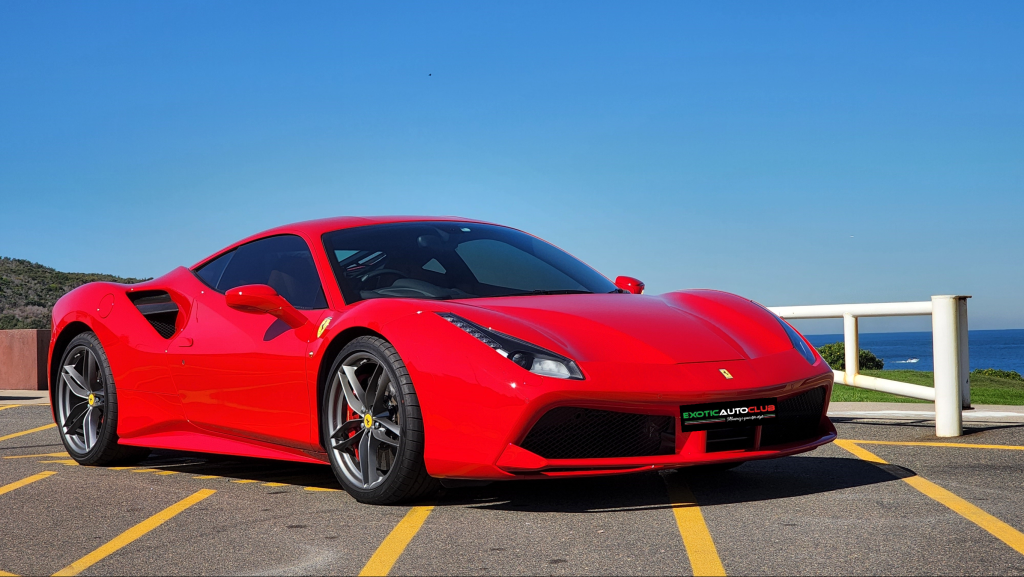 A sleek, red Ferrari 488 GTB with aerodynamic contours is parked in a lot with ocean views in the background. The car has dark tinted windows, black rims, and a low profile, capturing a sense of speed and elegance. The sky is clear and blue, enhancing the vibrant scene.