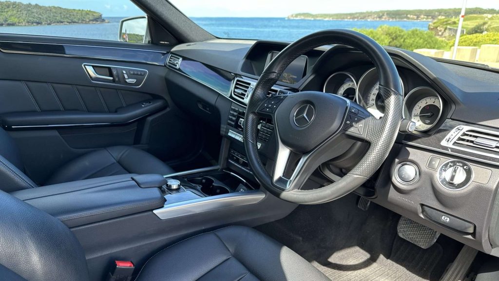 Interior view of a luxurious Mercedes-Benz E250 showcasing a sleek, leather-wrapped steering wheel, modern dashboard with various controls, leather seats, and a clear view through the driver's side window out toward a scenic coastline with blue water and greenery.