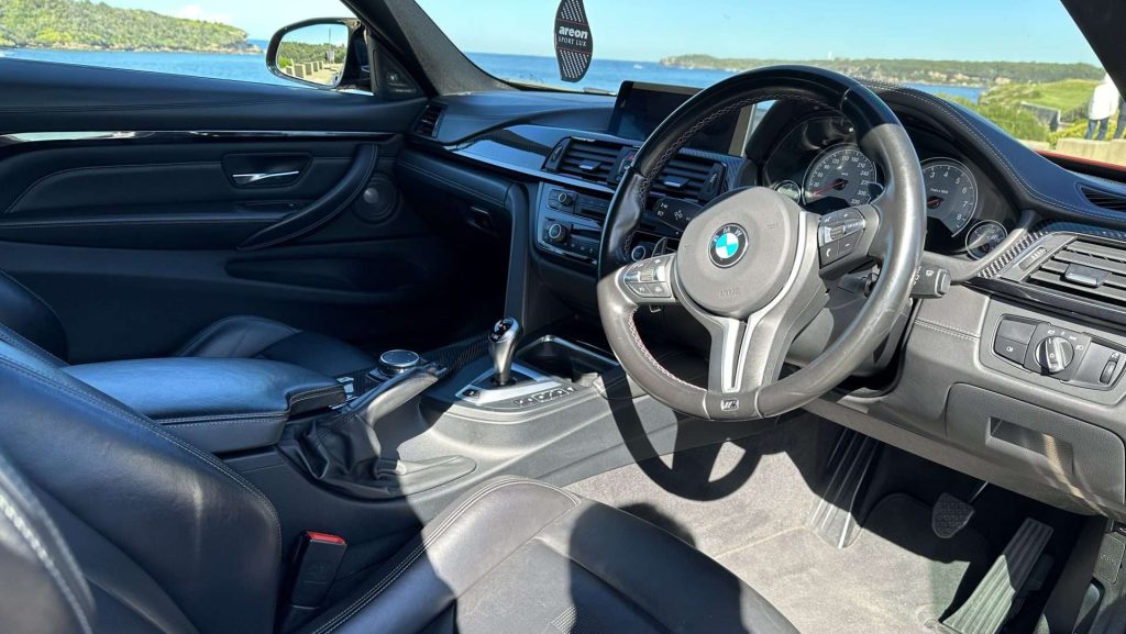 Interior view of a BMW M4, featuring a leather-wrapped steering wheel with the BMW logo, black leather seats, a gear shift, and a dashboard with various controls and a display screen. The car is parked near a scenic location with water and greenery visible through the windows.