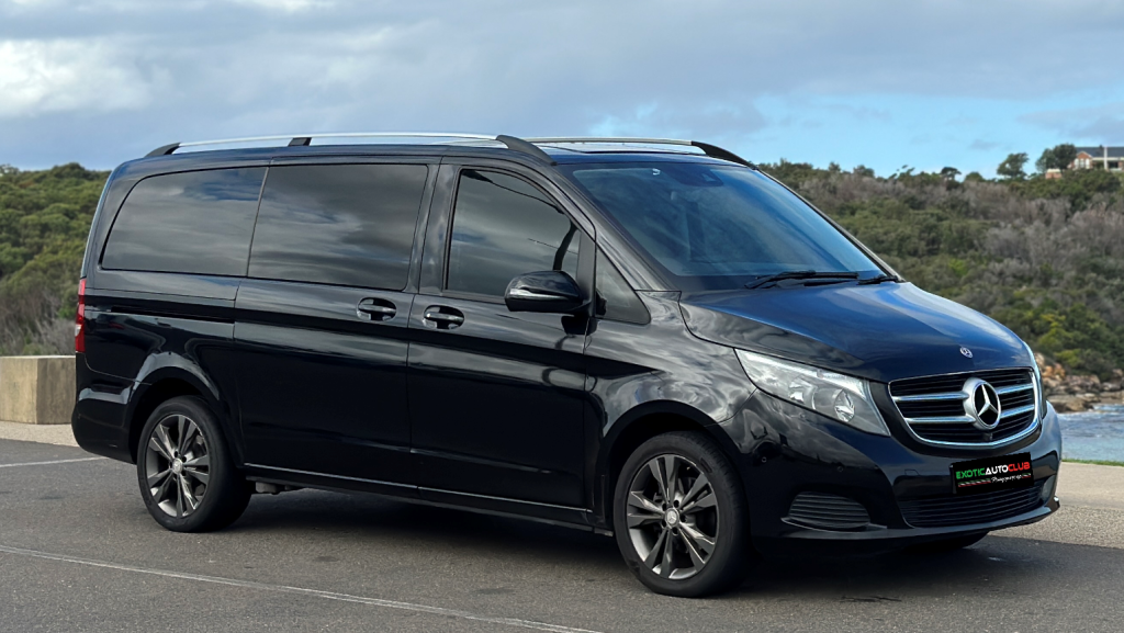A black Mercedes-Benz V220d van is parked on a roadside with a scenic background of trees and a cloudy sky. The van has tinted windows and a sleek design, and it is positioned to show its left side and front.