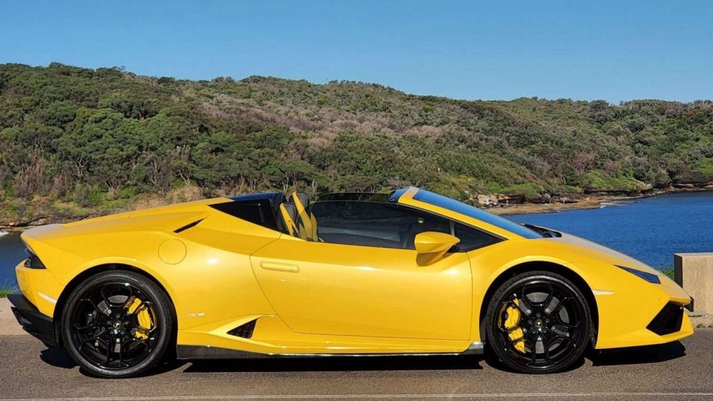 A yellow Lamborghini Huracan Spyder with a convertible top is parked in front of a scenic background featuring lush green trees and a body of water under a clear blue sky. The car has black wheels and yellow brake calipers.