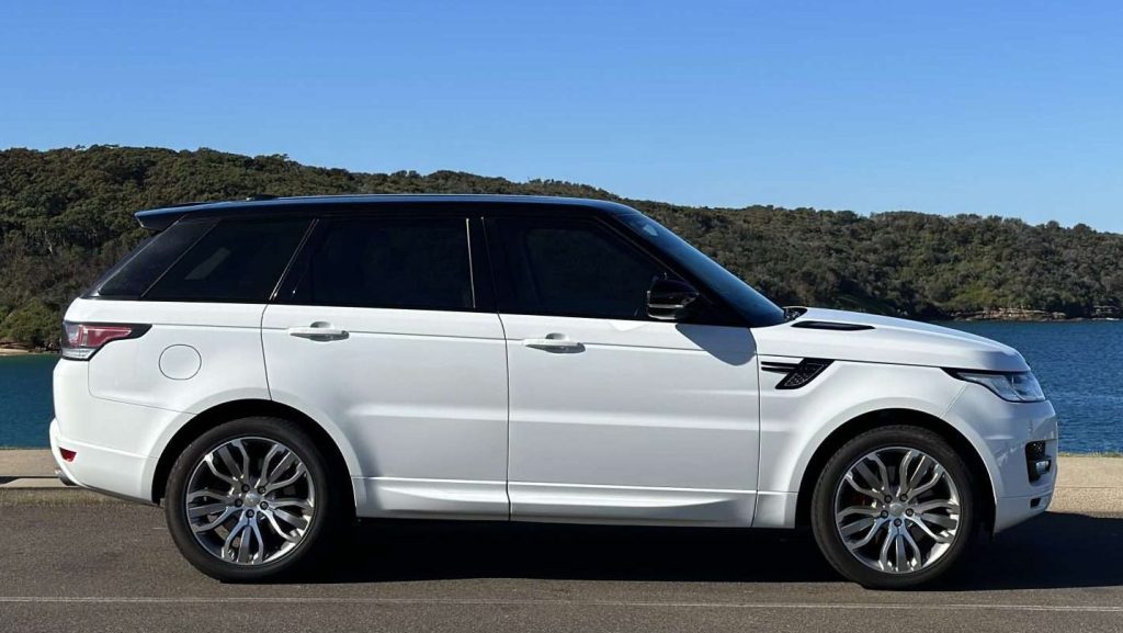 A sleek, white Range Rover Sport SUV is parked on a road with a scenic backdrop of a blue body of water and green, tree-covered hills under a clear blue sky. The vehicle is seen from a side profile view.