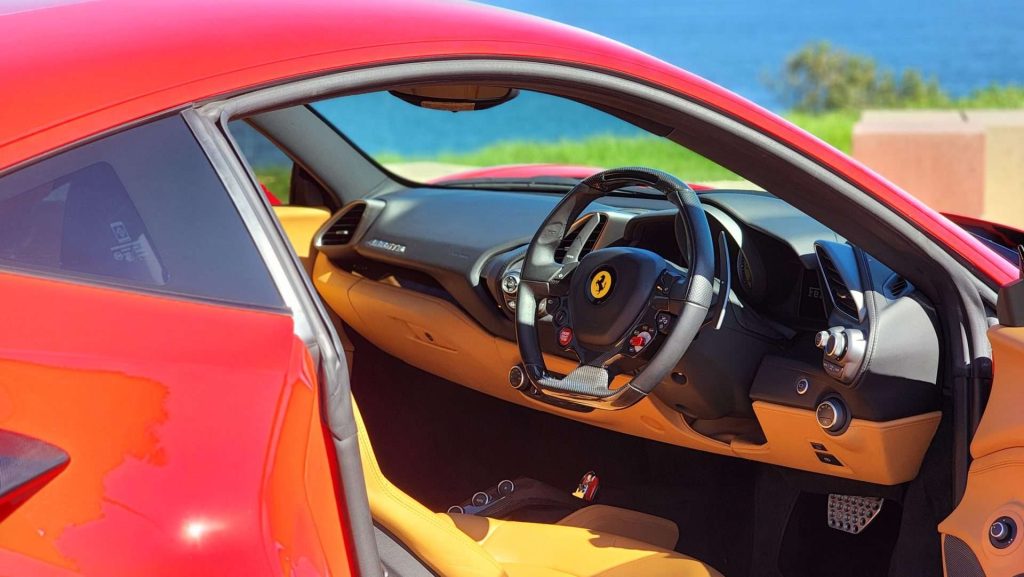 A vibrant red Ferrari 488 GTB with its driver-side door open reveals a luxurious tan leather interior and a sleek, modern dashboard. The steering wheel features a prominent black emblem with a yellow background. The car is parked near a scenic coastal view.