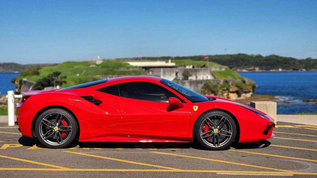 A sleek red Ferrari 488 GTB is parked on a paved area near a waterfront. The background features a body of water and a grassy, hilly area on the other side. The sports car stands out against the scenic backdrop under a clear blue sky.