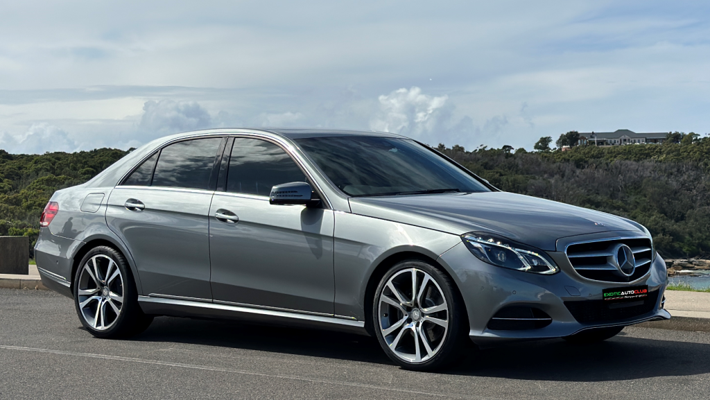 A sleek, silver Mercedes-Benz E250 sedan is parked on a paved surface against a backdrop of greenery and a cloudy sky. The car's design features smooth lines, tinted windows, and stylish alloy wheels, highlighting its luxurious aesthetics.