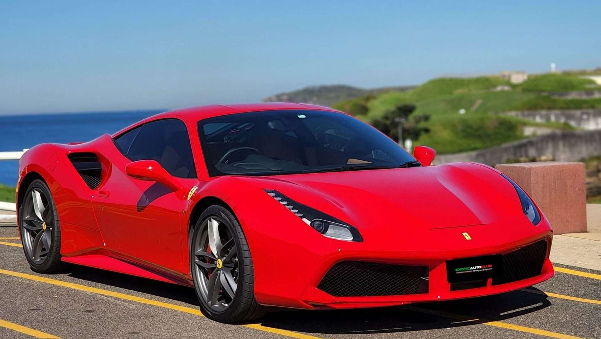 A bright red Ferrari 488 GTB is parked on a coastal road with the ocean and greenery in the background. The car boasts a sleek, aerodynamic design with sharp lines and distinctive headlights, showcasing its modern, high-performance build.