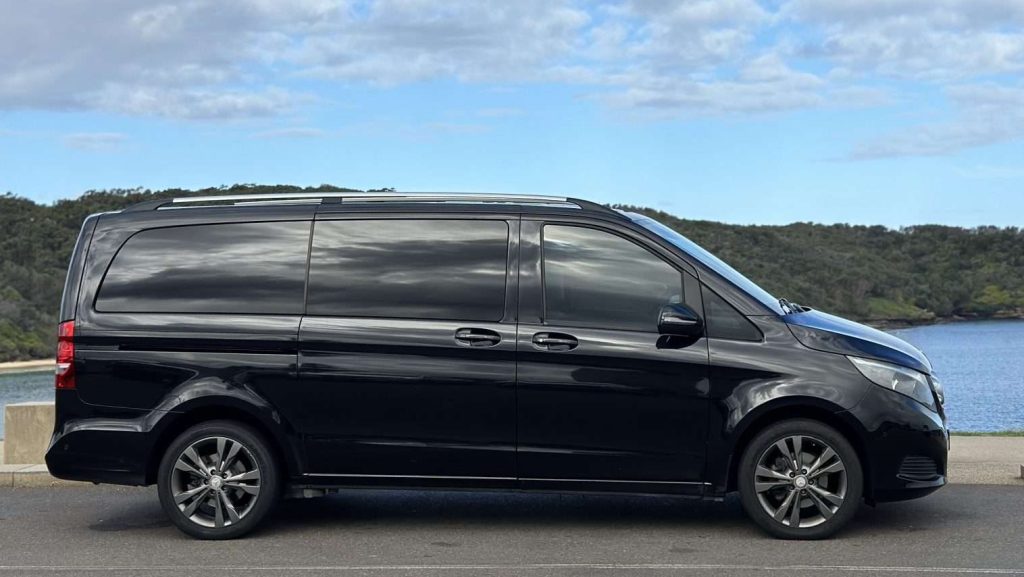 A black Mercedes-Benz V220d minivan is parked on a road adjacent to a body of water. The sky is partly cloudy, and there is a green, hilly landscape in the background. The van has tinted windows and dark alloy wheels.