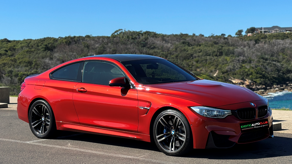 A shiny red BMW M4 sports car is parked in an outdoor setting with lush greenery and a clear blue sky in the background. The car has black rims, tinted windows, and a black roof. The scenery includes a body of water and trees in the distance.
