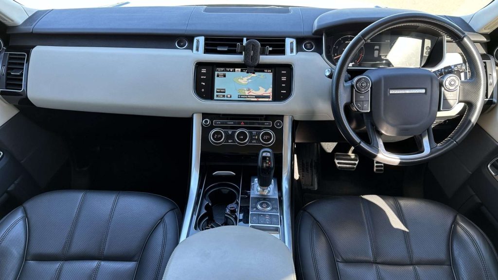 The interior of the Range Rover Sport luxury car features a touchscreen infotainment system displaying a map, a multifunction steering wheel, digital and analog gauges, leather seats, and a central console with various controls and gear lever.