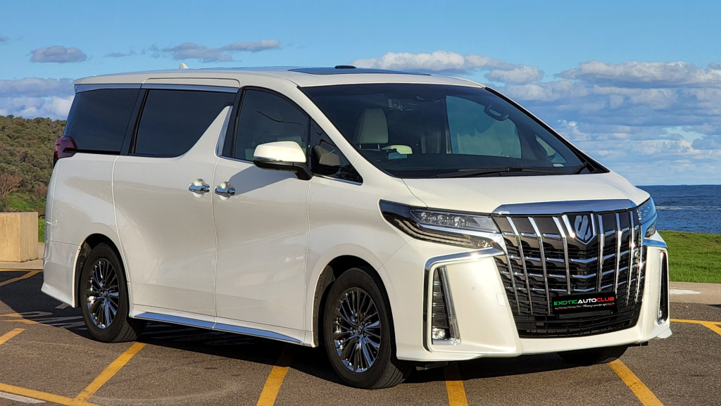 A white luxury S Alphard minivan parked on a road near a grassy landscape and a body of water under a blue sky with scattered clouds. The executive lounge features a sleek design with a prominent front grille, shiny alloy wheels, and tinted windows.