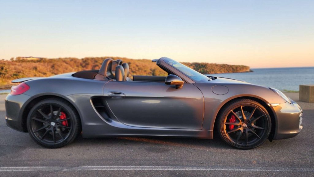 A sleek gray Porsche Boxster S convertible with the top down, parked by a scenic coastal area with trees and water in the background. The car has red brake calipers and black alloy wheels. The sun is setting, casting a warm light over the scene.