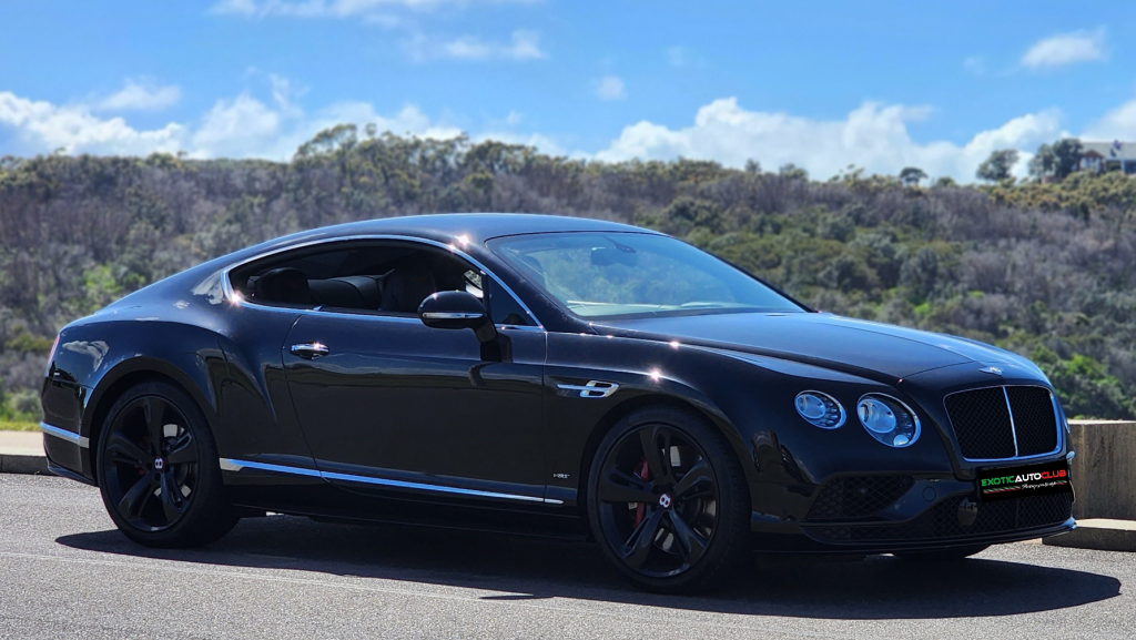 A sleek black Bentley Continental GT luxury coupe is parked in a scenic outdoor setting with trees and bushes in the background. The car has a modern design with tinted windows and black rims, and the sky above is blue with scattered clouds.