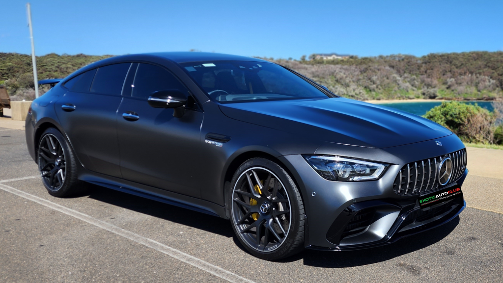 A sleek, dark gray Mercedes-Benz AMG GT63s is parked on a sunlit road near a body of water with green hills in the background. The car features tinted windows, large black alloy wheels with yellow brake calipers, and a stylish front grille.