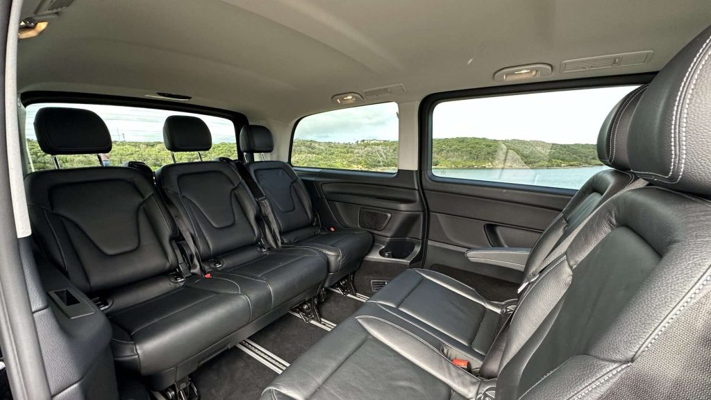 Interior of a spacious Mercedes-Benz V220d with three rows of black leather seats. The picture is taken from the passenger side, showing the rear and middle rows. Large windows provide a view of greenery outside. The interior appears clean and modern.