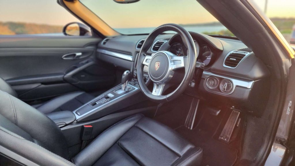Interior of a luxury Porsche Boxster S convertible sports car with black leather seats. The focus is on the sleek design and modern dashboard featuring a central touchscreen, multifunction steering wheel, and an automatic gear shift. The car is parked near a scenic outdoor background.