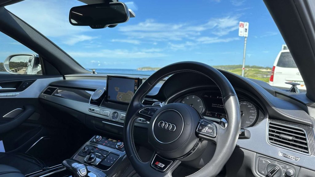 The interior of an Audi S8 is shown, featuring the steering wheel, dashboard, and infotainment system. The car is parked near a coastal area, with a view of the ocean and a clear blue sky through the windshield. A white van is visible in the background.