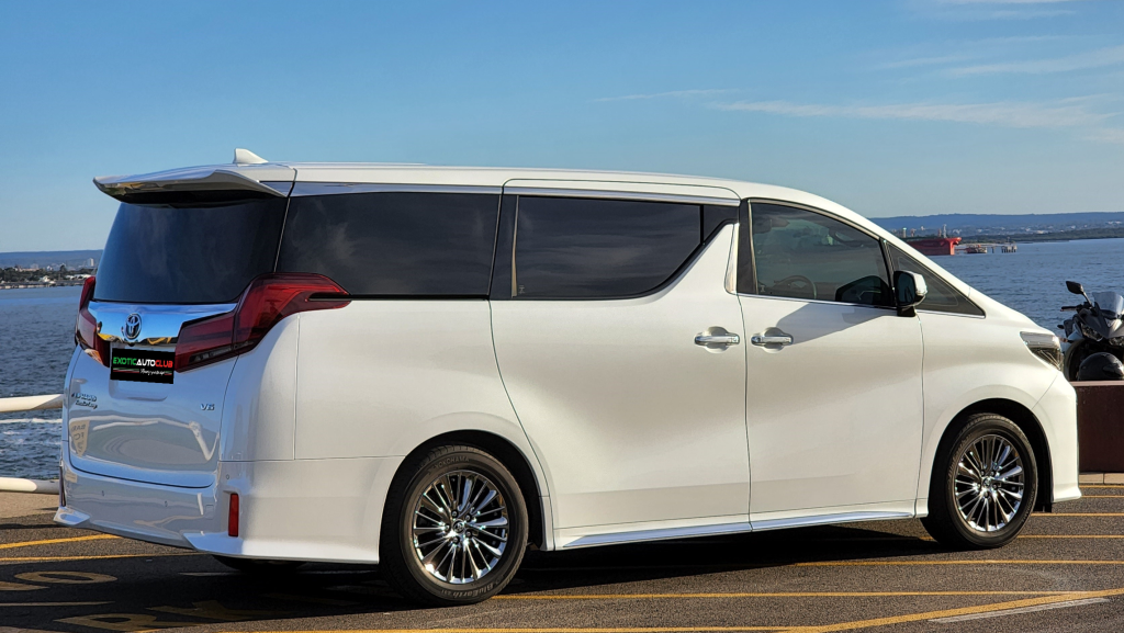 A white Alphard minivan parked by the waterfront on a sunny day. The executive lounge features tinted windows and shiny alloy wheels, with a body of water and a distant shoreline under a clear blue sky in the background.