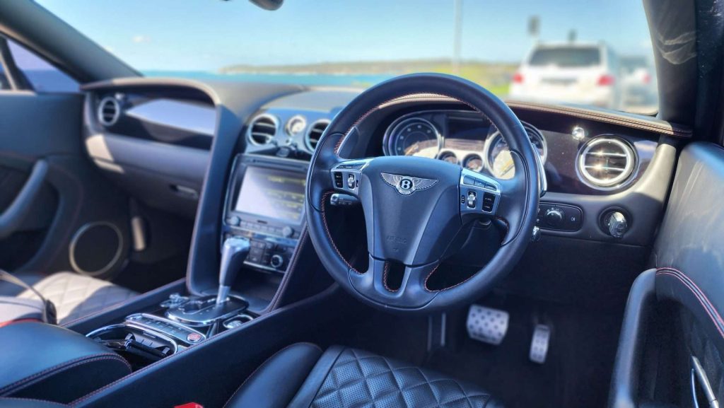 Close-up of a Bentley Continental GT's interior featuring a leather-wrapped steering wheel with the Bentley logo, a dashboard with analog dials, a central touchscreen display, and quilted leather seats. A white vehicle is visible through the windshield in the background, showcasing the V8's elegance.