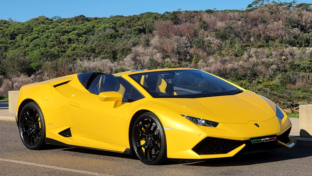 A yellow Lamborghini Huracan Spyder convertible is parked on a paved road with a scenic backdrop of trees and hills. The luxury sports car has a sleek, aerodynamic design and black alloy wheels. The road is wide, and the skies are clear, enhancing the vibrant color of the car.