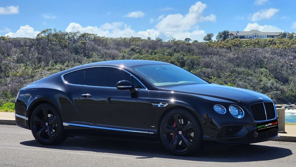 A sleek black Bentley Continental GT V8s is parked on the side of a road with lush green hills and a clear blue sky in the background. The luxury car features tinted windows, alloy wheels, and the distinctive Bentley grille.