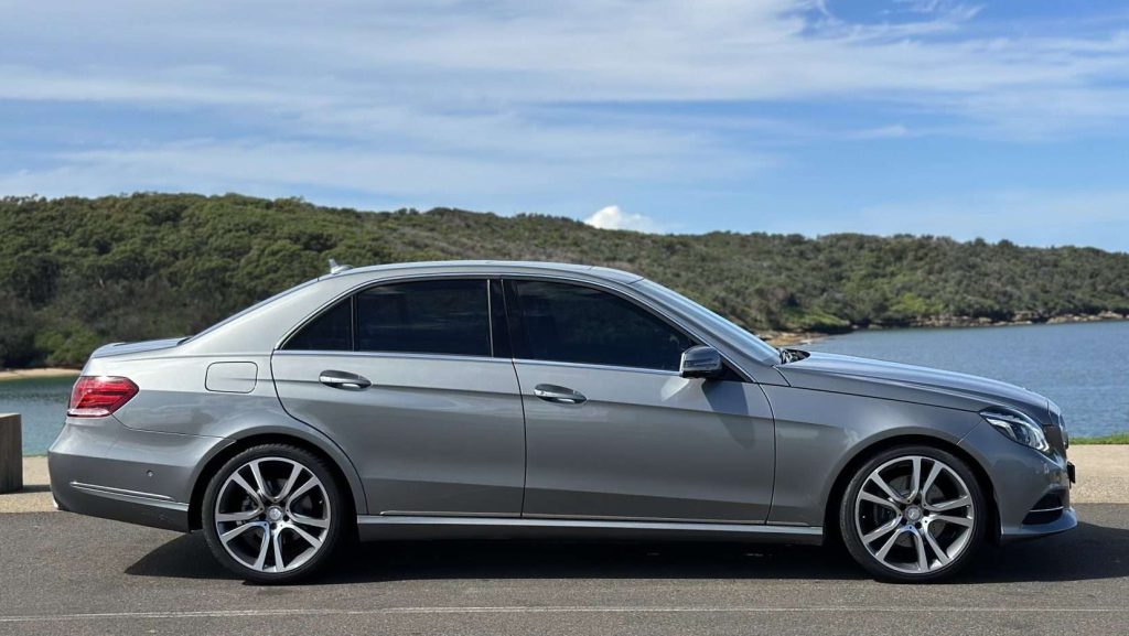 A Mercedes-Benz E250 with tinted windows is parked on a road near a water body, surrounded by lush greenery and a partly cloudy sky. The silver luxury car features sleek lines and alloy wheels, giving it a modern and stylish appearance.