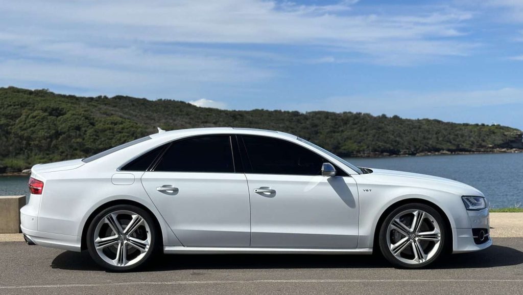 A white luxury Audi S8 sedan is parked on a road with a scenic backdrop of greenery, water, and a partly cloudy sky. The car has dark tinted windows and large alloy wheels.