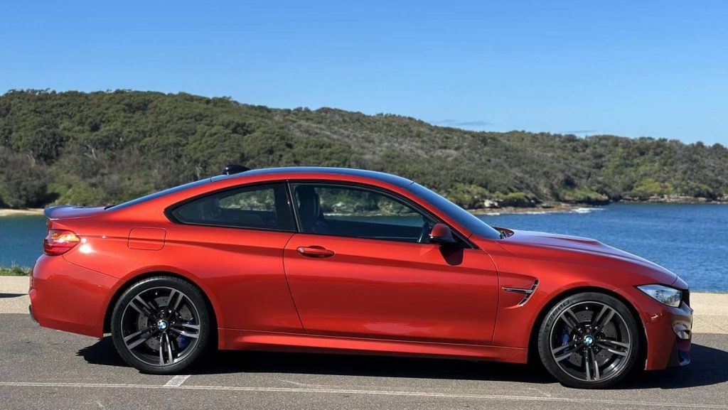 A red BMW M4 with black rims is parked on a paved surface near a body of water. In the background, there's a lush green hillside under a clear blue sky. The car's side profile highlights its sleek design and aerodynamic features.