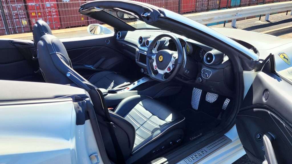 A sleek and modern Ferrari California convertible sports car with a gray exterior and a luxurious black leather interior. The driver's seat and dashboard are visible, featuring a sporty steering wheel with the iconic Ferrari logo and an array of controls.