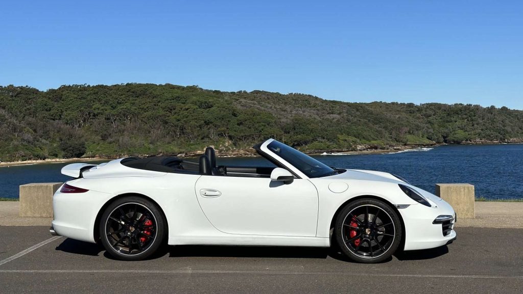 A white Porsche 911 Carrera S cabrio is parked on a paved surface with its top down. In the background, there is a scenic view of a forest and a body of water under a clear blue sky.