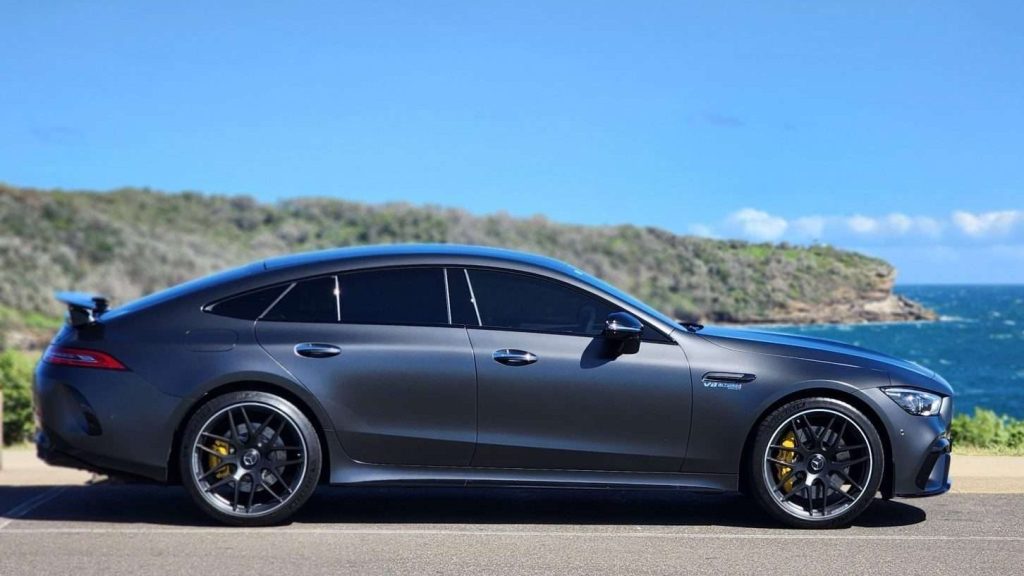 A matte grey Mercedes-AMG GT63s four-door coupe with yellow brake calipers is parked on a coastal road. The car is in side profile view, with a scenic background of rocky cliffs, greenery, and the ocean under a clear blue sky.