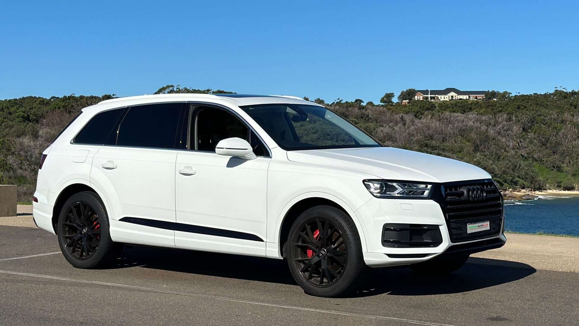 A white Audi SUV with black alloy wheels is parked on a paved area near a body of water. The background features green hills and sparse houses under a clear blue sky, making it look like it's ready to compete in the V8SC sport circuit.