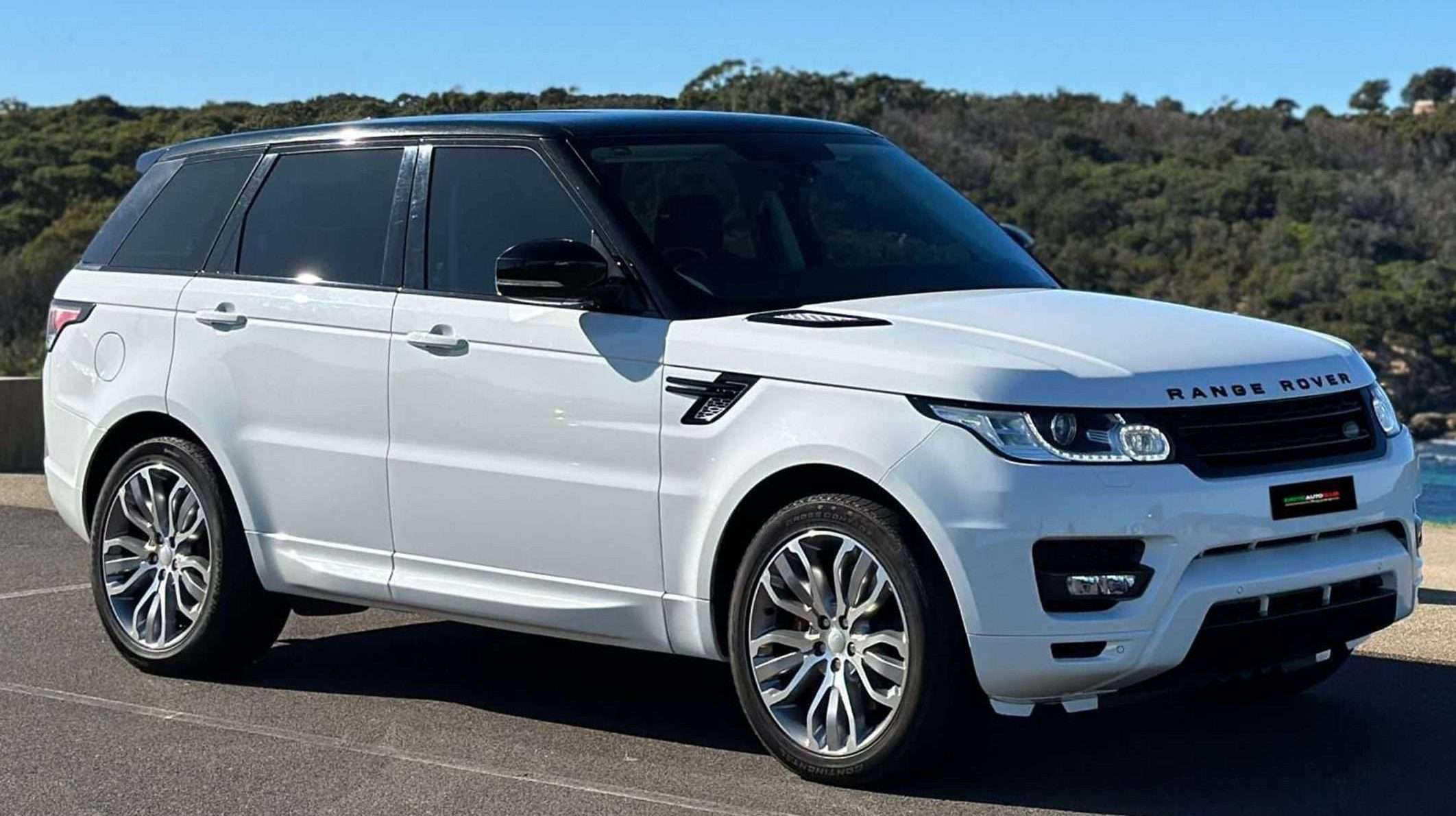 A white Range Rover Sport SUV is parked on a sunny day against a scenic backdrop of trees and a body of water. The vehicle features a black roof, tinted windows, and silver wheels. The background shows a clear blue sky, enhancing the overall view.