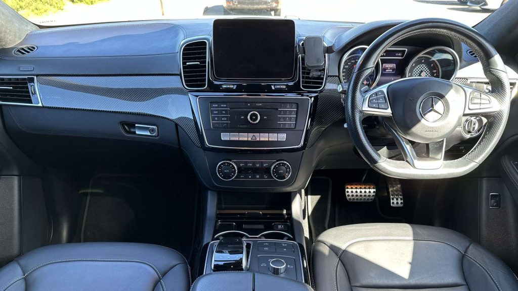 The image shows the interior of a modern Mercedes-Benz, specifically the GLE350d Orange Art Edition. The dashboard includes a touchscreen display, central control panel with various buttons, air conditioning controls, and a steering wheel with multiple buttons. The seats are made of leather.