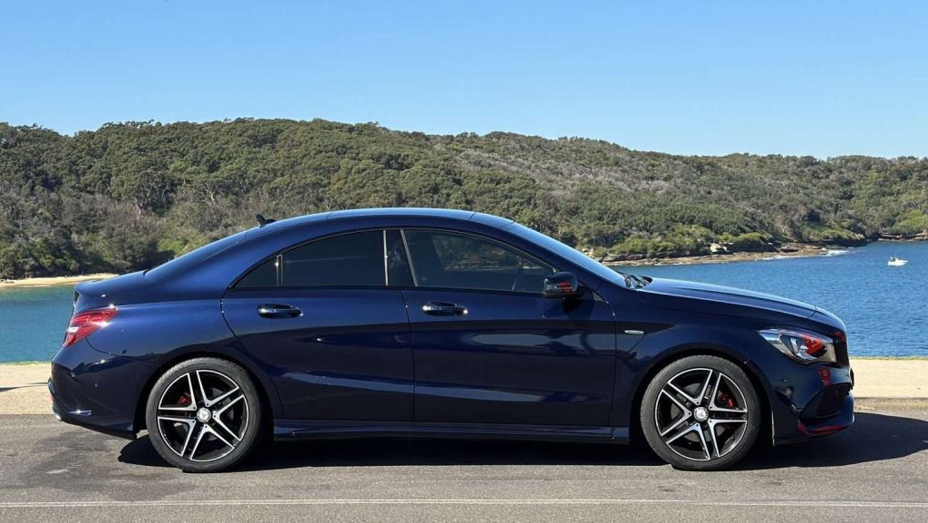 A sleek, dark blue Mercedes-Benz CLA250 is parked in front of a scenic view, featuring a serene body of water and lush green trees in the background. The luxury car has distinctive alloy wheels and tinted windows, and the scene is bathed in clear, sunny weather.