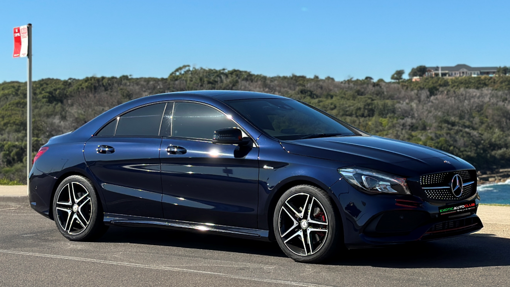 A sleek, dark blue Mercedes-Benz CLA250 sedan is parked on a paved surface with coastal scenery in the background. The car features tinted windows and distinctive alloy wheels, set against a backdrop of greenery and a clear blue sky.