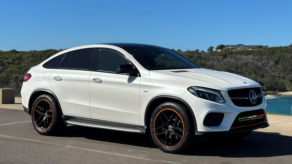 A white Mercedes-Benz GLE350d coupe, part of the sleek Orange Art Edition, is parked by a scenic coastal area under a clear blue sky. The SUV boasts black wheels with bold orange accents, tinted windows, and a modern design. Dense green foliage and a distant house enhance the picturesque backdrop.