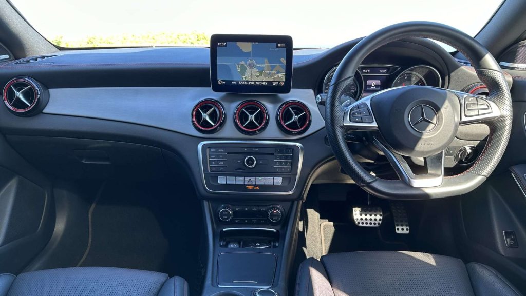 The interior of the Mercedes-Benz CLA250 focuses on the dashboard and steering wheel. The dashboard features a central infotainment screen displaying a map, three round air vents, various control buttons, and dials. The steering wheel boasts multiple buttons and logos.