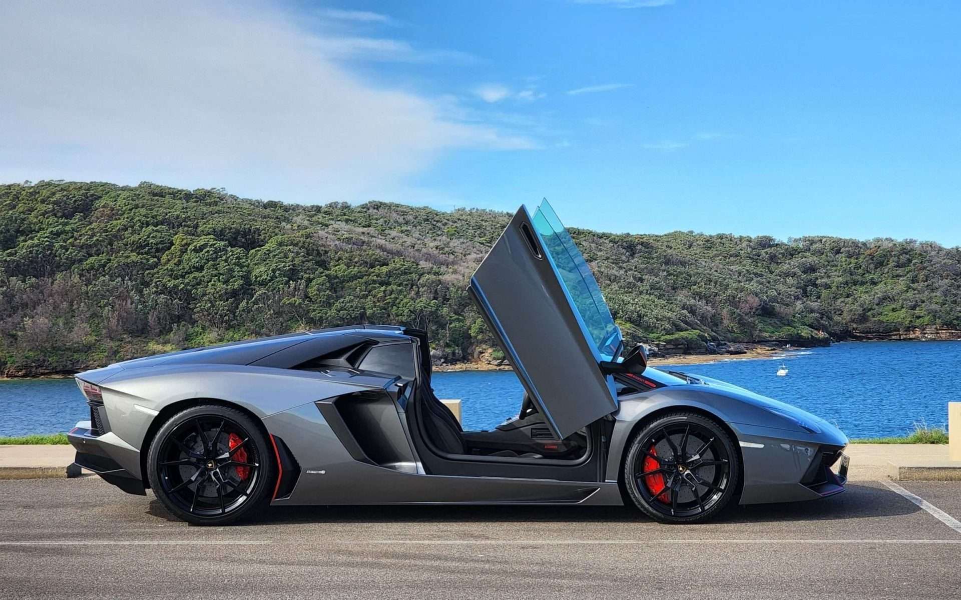 A sleek, silver luxury sports car with its futuristic scissor doors open, parked near a picturesque waterfront. Available through our lease program, the car features a stylish aerodynamic design with black rims and red brake calipers, set against the serene blue lake and lush green hills.