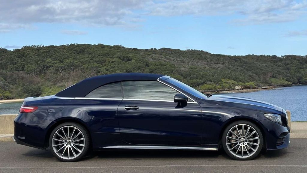 A sleek, dark blue Mercedes-Benz Cabrio with its top up is parked near a body of water. The background features a lush, green tree line under a partly cloudy sky, contributing to a serene and picturesque setting.