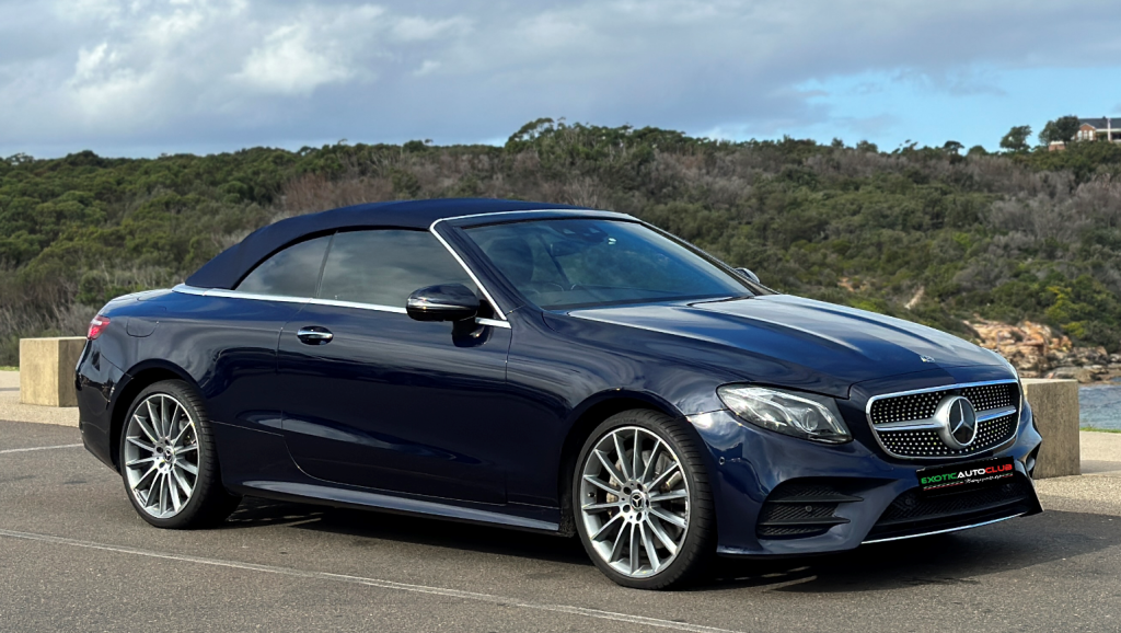 A sleek, dark blue Mercedes-Benz E300 Cabrio is parked on a paved area near a coastal scene with green trees and bushes in the background. The car has a black soft top and silver alloy wheels, with cloudy skies overhead.