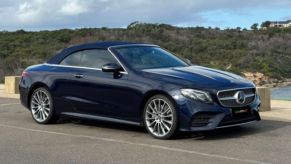 A dark blue Mercedes-Benz E300 Cabrio is parked on a paved surface near a scenic coastal area. The car features a sleek design, alloy wheels, and a soft top roof. Bushes and a body of water are visible in the background under a cloudy blue sky.