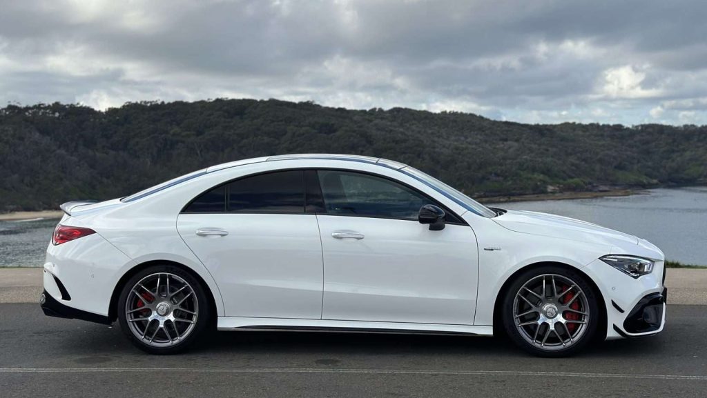 A white, sporty Mercedes-Benz Cla45s sedan is parked on a scenic roadway with a backdrop of lush greenery and a body of water. The car features sleek lines and red brake calipers, indicative of its AMG high-performance design. The sky is overcast with clouds.
